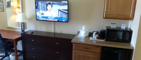 A hotel room with a television displaying a news program, a desk with a lamp, a microwave, and a cabinet.