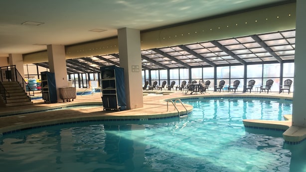 An indoor swimming pool with clear water, surrounded by lounge chairs and tables, and a high ceiling with skylights.