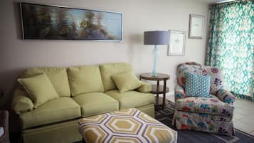 A living room with a green sofa, a patterned armchair, a small round table with a lamp, and a painting on the wall.