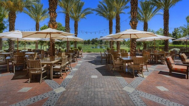An outdoor patio area with wooden tables and chairs, surrounded by palm trees.