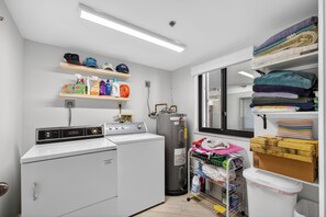 A laundry room with a front-loading washer and dryer, shelves with cleaning supplies, a water heater, and a window.