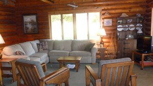 A cozy log cabin interior with a sectional sofa, wooden chairs, a coffee table, a television, and a cabinet with dishes.