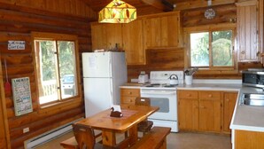 A wooden cabin kitchen with a refrigerator, stove, and wooden cabinets. There is a table with benches and a hanging light fixture.