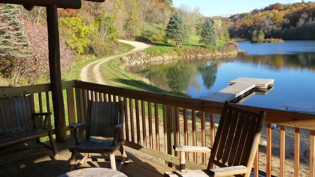 A wooden deck with two chairs and a table overlooking a calm lake, surrounded by trees and a dirt path.
