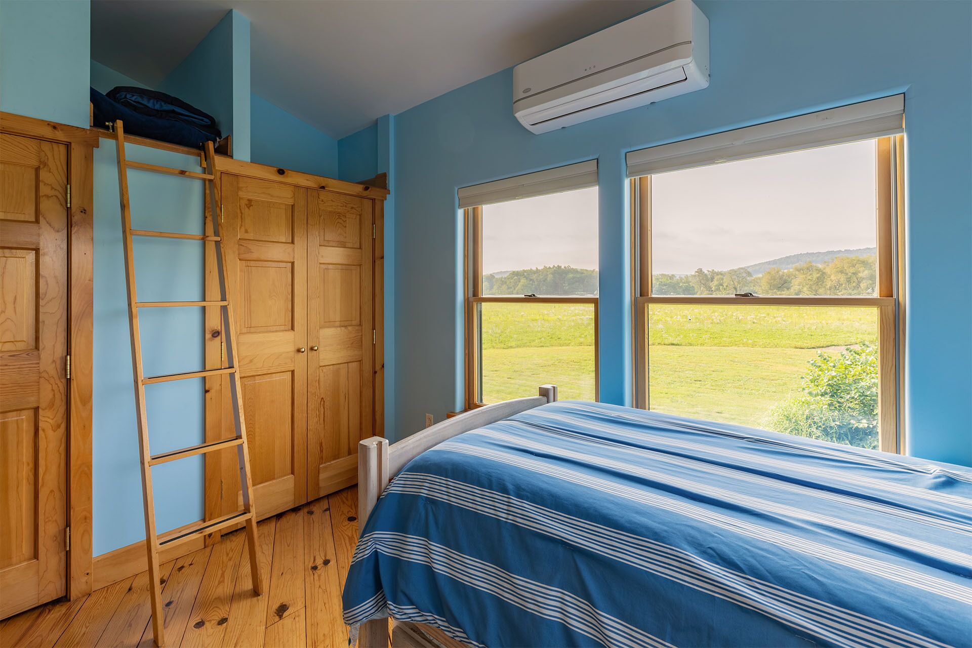 Une chambre avec un lit superposé, une échelle en bois et une grande fenêtre donnant sur un champ verdoyant et des collines.