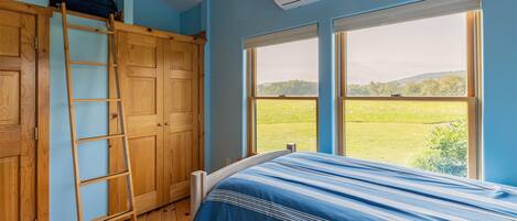 Une chambre avec un lit superposé, une échelle en bois et une grande fenêtre donnant sur un champ verdoyant et des collines.