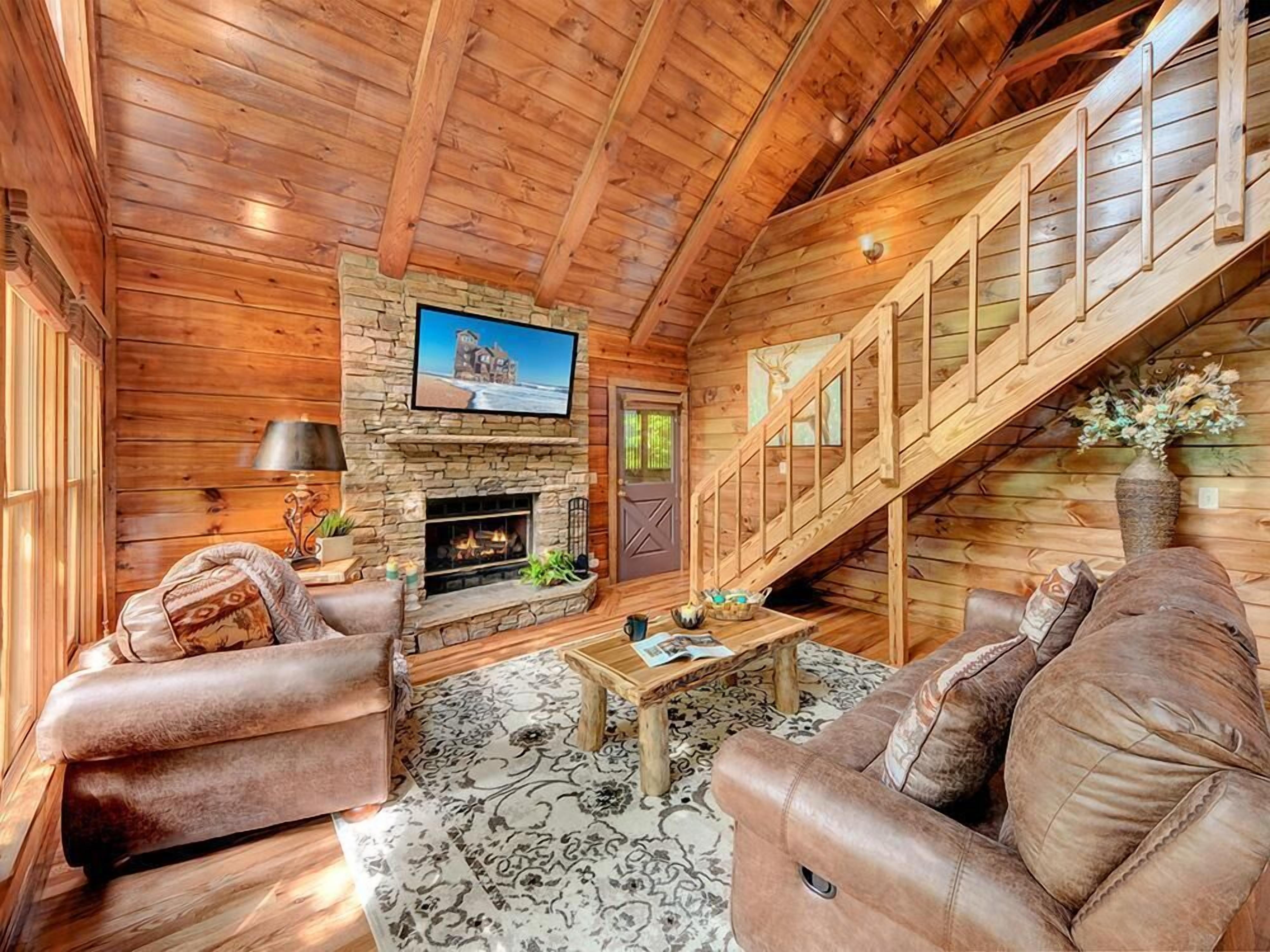 L'intérieur d'une cabane en rondins, avec une cheminée en pierre, un escalier en bois, des canapés en cuir et un téléviseur fixé au-dessus de la cheminée.