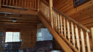 Interior de una cabaña de madera con escalera de madera, un sillón de cuero, una mesa de comedor y un refrigerador.