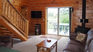 Interior de una cabaña de madera con escalera, televisor, chimenea y puerta corrediza de vidrio que da a un espacio exterior.