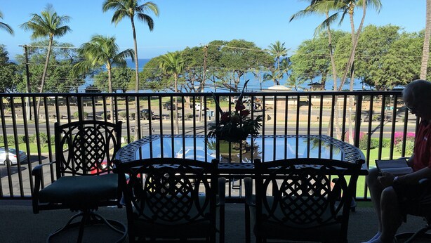 A balcony with a round table and two chairs, overlooking a street with palm trees and a view of the ocean. - Cozy Top-Floor  Maui Parkshore Condo (Kihei)
