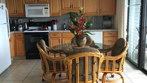 A kitchen with wooden cabinets, a white microwave, a stove, and a dining table with wicker chairs. - Cozy Top-Floor  Maui Parkshore Condo (Kihei)