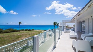 A balcony with white chairs overlooking a beach and ocean, featuring a clear sky and distant airplane. - Stunning Penthouse on Mullet Bay beach (Cupecoy)