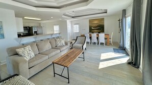 A spacious living area with a sofa, coffee table, and dining table. A kitchen is visible in the background. - New Renovation 2024 Direct Gulf View! White Beach Dolphins 4 Bedrooms, 3.5 bath (Gulf Shores)