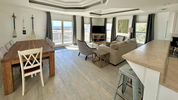 A spacious living area with a wooden dining table, a sofa, and a TV. - New Renovation 2024 Direct Gulf View! White Beach Dolphins 4 Bedrooms, 3.5 bath (Gulf Shores)