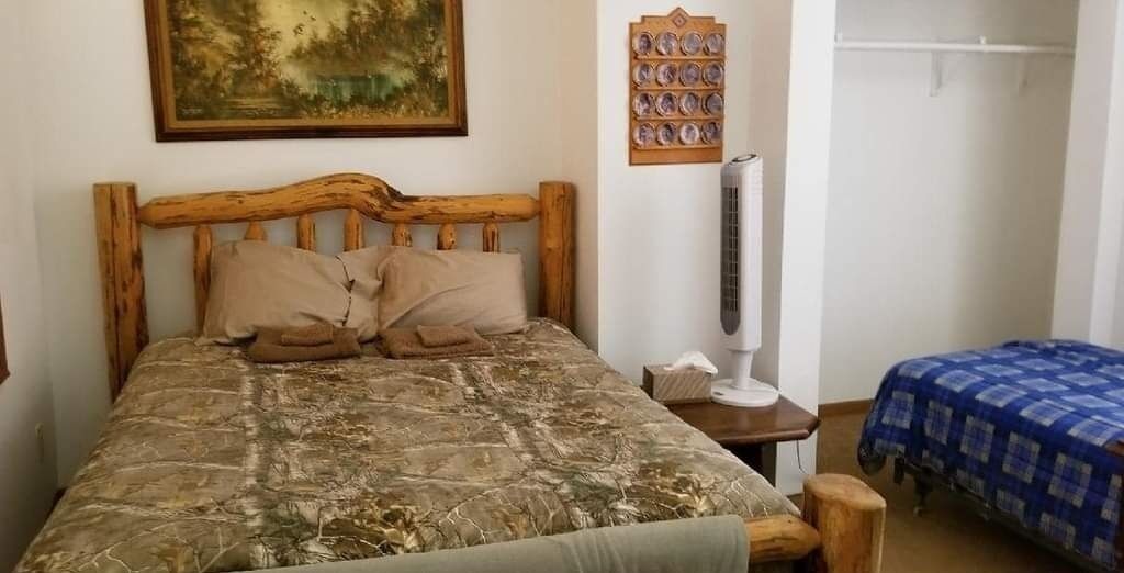 Un dormitorio con cabecera de madera, una cama con cubrecama estampado camuflaje, una mesita de noche con una lámpara y una colcha azul en una cama más pequeña.