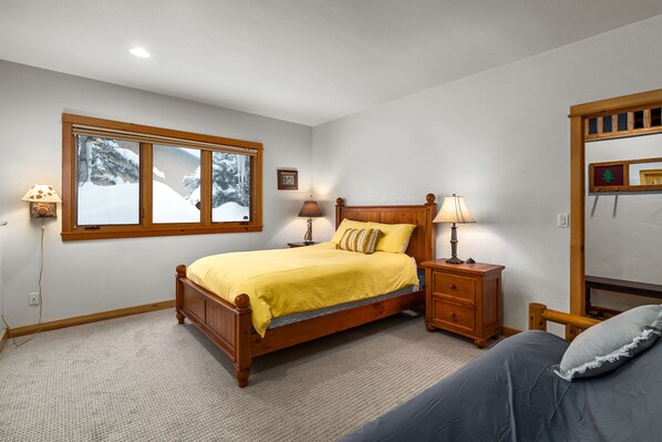 Une chambre à coucher avec un lit en bois, deux tables de chevet avec des lampes, une fenêtre donnant sur des arbres couverts de neige et un miroir avec un cadre en bois.