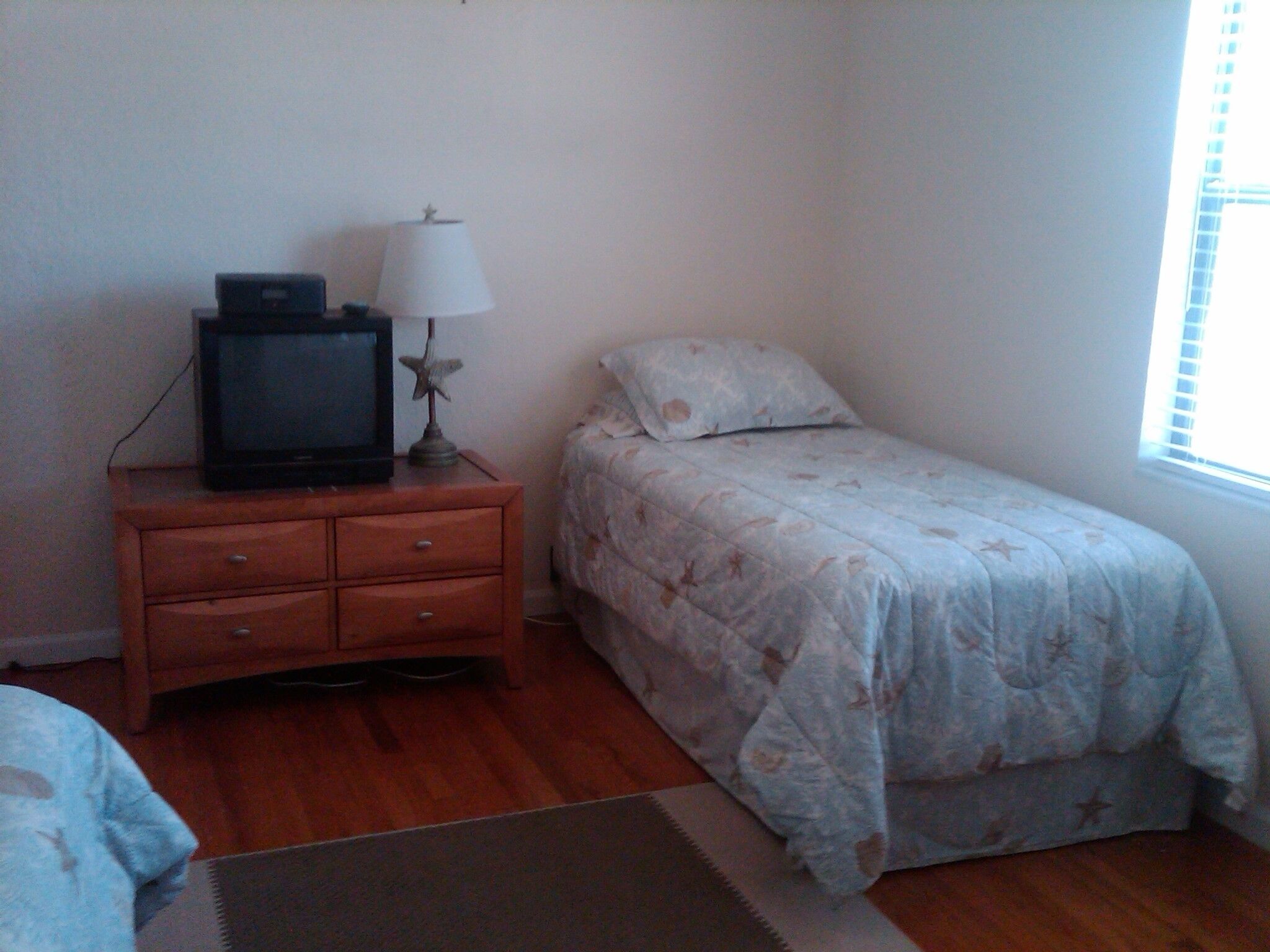 A bedroom with a bed, a nightstand with a television and lamp, and a window with blinds.
