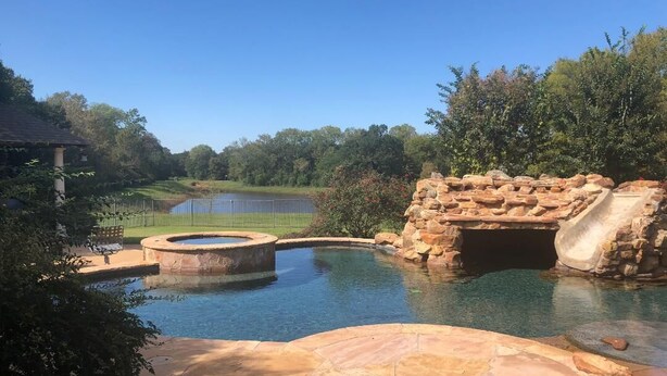 A swimming pool with a stone bridge and a small waterfall, surrounded by greenery and a distant body of water. - Whitetail Ranch Manor Retreat (Bryan)
