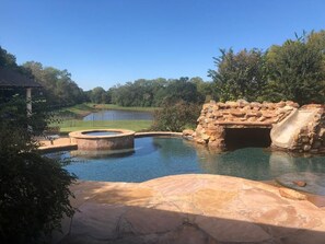 A swimming pool with a stone bridge and a small waterfall, surrounded by greenery and a distant body of water. - Whitetail Ranch Manor Retreat (Bryan)