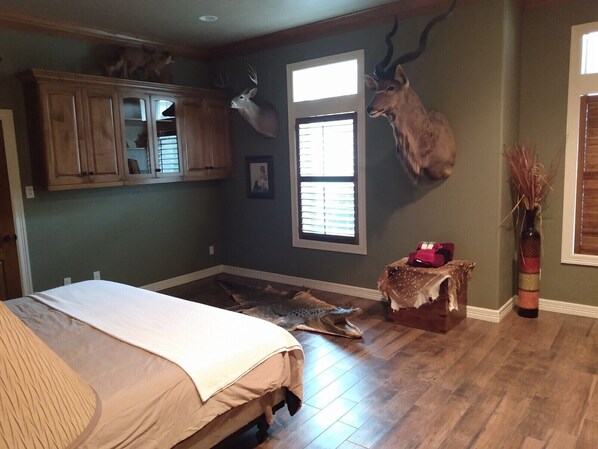 A room with a bed, wooden cabinets, a deer head mounted on the wall, and a window with blinds. - Whitetail Ranch Manor Retreat (Bryan)