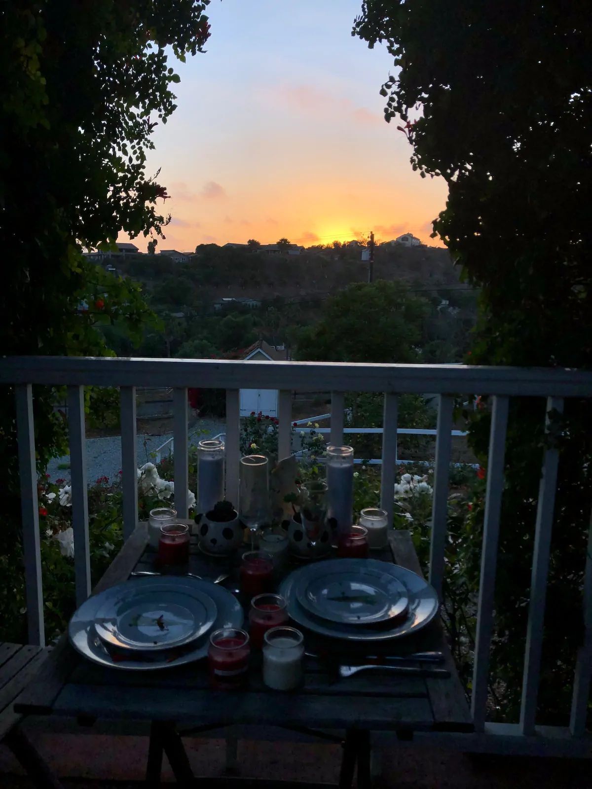 Mesa al aire libre con platos, velas y un tocadiscos de vinilo, con vistas a un paisaje panorámico de una puesta de sol y edificios al fondo.