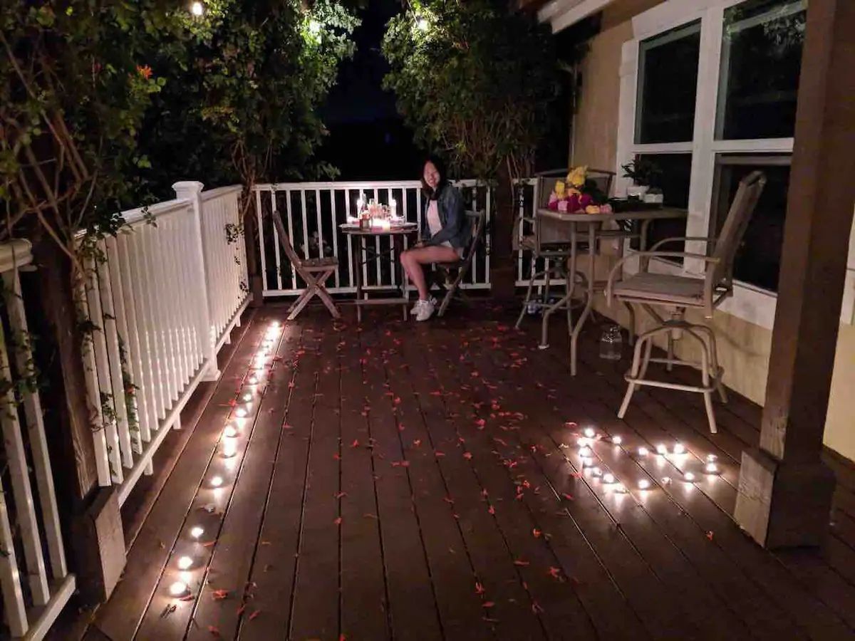 A wooden deck with string lights and scattered rose petals, a white fence, and a table with chairs.
