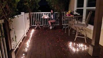 A wooden deck with string lights and scattered rose petals, a white fence, and a table with chairs.