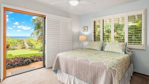A bedroom with a bed, a ceiling fan, a window with shutters, and a view of a garden and ocean. - Luxury Oceanfront Escape – A/C, Hot Tub, 5 bedrooms & Stunning Ocean Views! (Princeville)