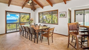 A dining area with a wooden table and chairs, large windows with views of palm trees and the ocean, and a ceiling fan. - Luxury Oceanfront Escape – A/C, Hot Tub, 5 bedrooms & Stunning Ocean Views! (Princeville)