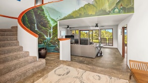 A modern interior with a curved staircase, a living area with a kitchen, and a mural of a mountainous landscape. - Luxury Oceanfront Escape – A/C, Hot Tub, 5 bedrooms & Stunning Ocean Views! (Princeville)