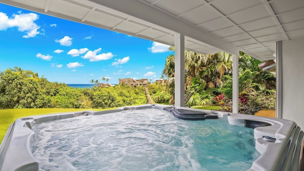 A hot tub with a view of a lush garden and a distant building. - Luxury Oceanfront Escape – A/C, Hot Tub, 5 bedrooms & Stunning Ocean Views! (Princeville)