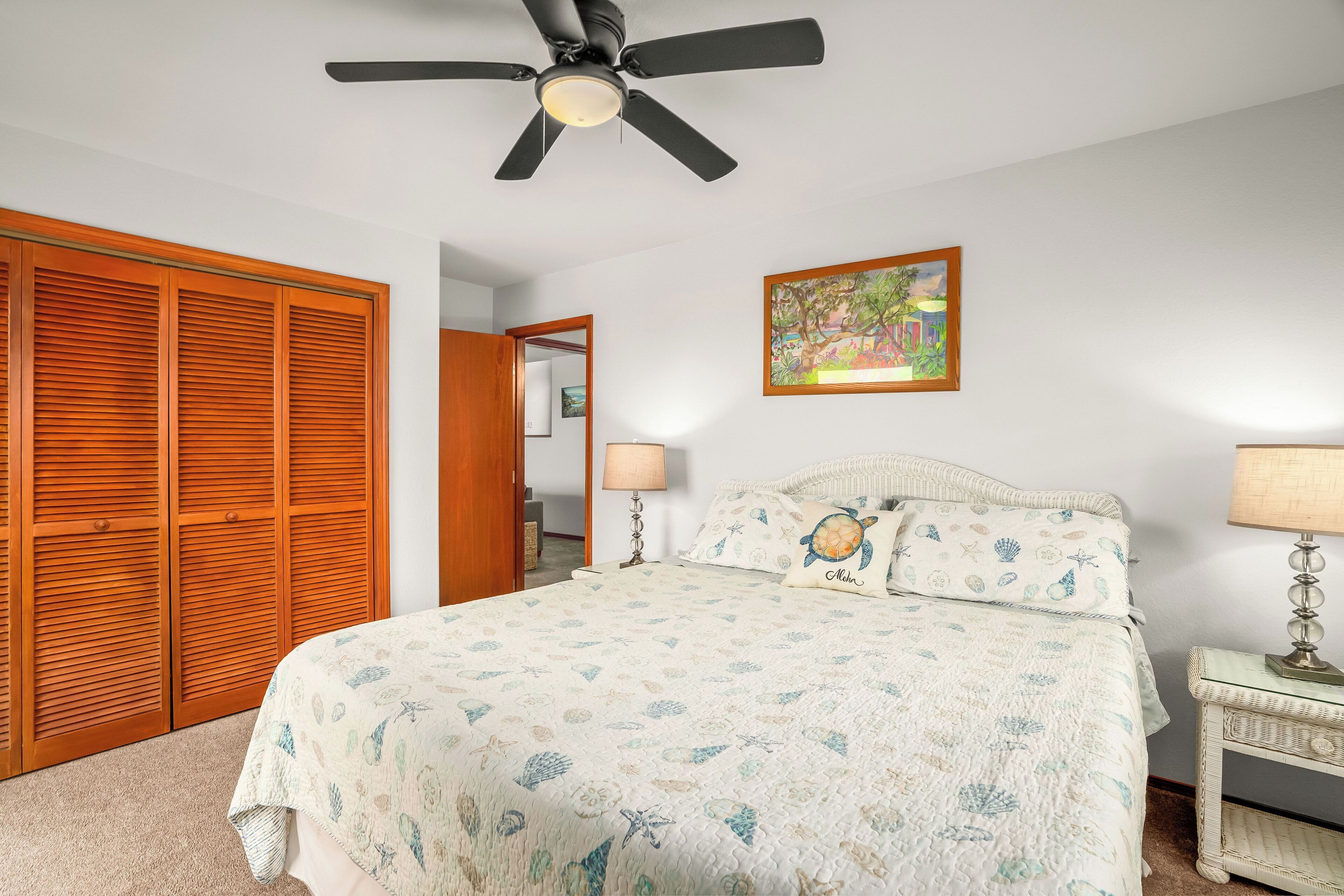 Un dormitorio con cama, ventilador de techo, mesita de noche con lámpara, armario de madera y un cuadro en la pared.