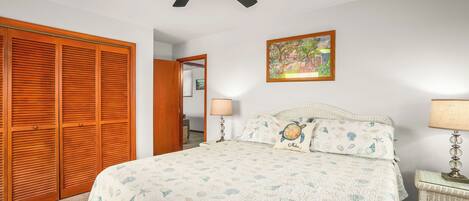 Un dormitorio con cama, ventilador de techo, mesita de noche con lámpara, armario de madera y un cuadro en la pared.