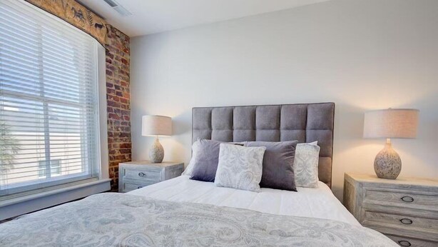 Quarto com uma cama grande, duas mesas de cabeceira, parede de tijolos e uma janela com persianas.