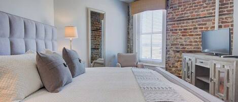 A hotel room with a large bed, a television, a brick wall, and a window with blinds.