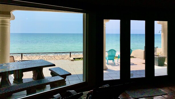A room with a view of the sea through a large window, a table, and chairs outside.