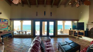 A spacious living room with a large TV, leather sofas, and a view of the sea through large glass doors.