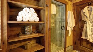 A wooden bathroom with shelves holding rolled towels, a glass shower enclosure, and a robe hanging on the door. - Refined creekside retreat • Barrel sauna overlooking the river • Hot tub • Starlight evenings • Minutes to Sundance (Sundance)