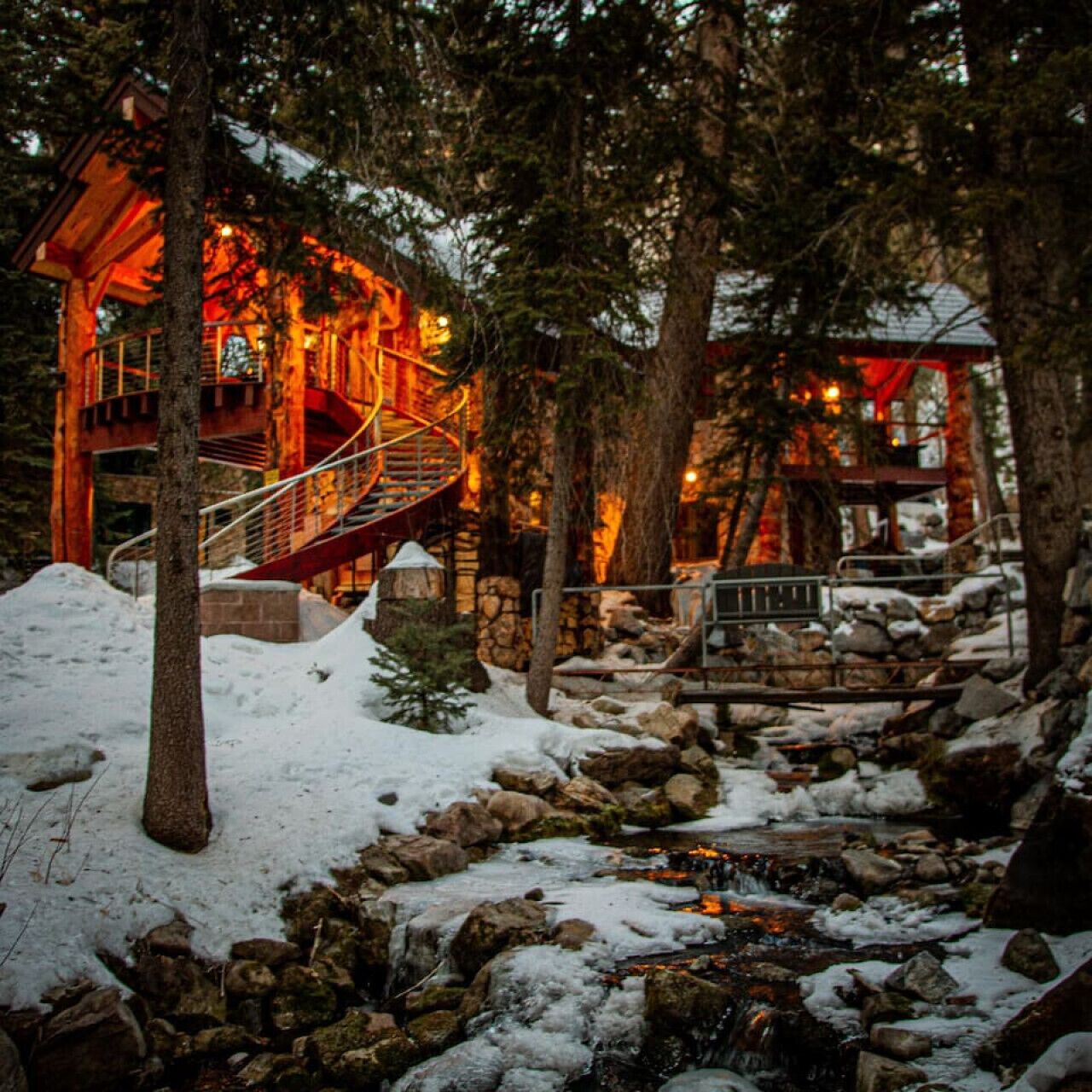 Una baita con una scala di legno che conduce a un livello superiore illuminato di rosso, circondata da neve e alberi.