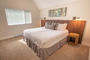 Een slaapkamer met een groot bed, een hoofdbord, een rond nachtkastje, een raam met jaloezieën en een schilderij aan de muur.