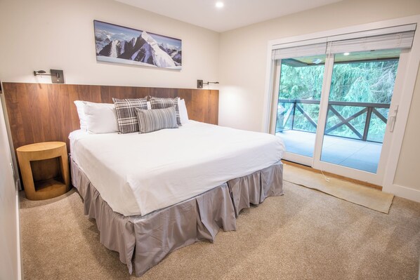 A bedroom with a large bed, a wooden headboard, a small side table, a large window with a view of trees, and a painting on the wall.