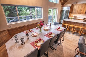 A dining area with a long table set for a meal, wooden flooring, and large windows overlooking a forested area. - Pinnacle Ridge 23 - Ski in Ski out, Private Hot Tub, Gas Fireplace (Whistler)