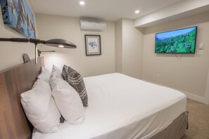 A neatly made bed with white and plaid pillows, a mounted TV displaying a nature scene, and an air conditioning unit on the wall.