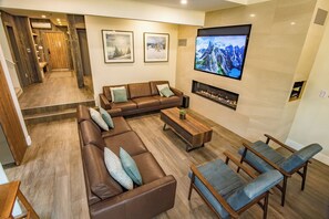 A modern living room with a brown leather sofa, blue armchairs, a wooden coffee table, and a flat-screen TV mounted on the wall.