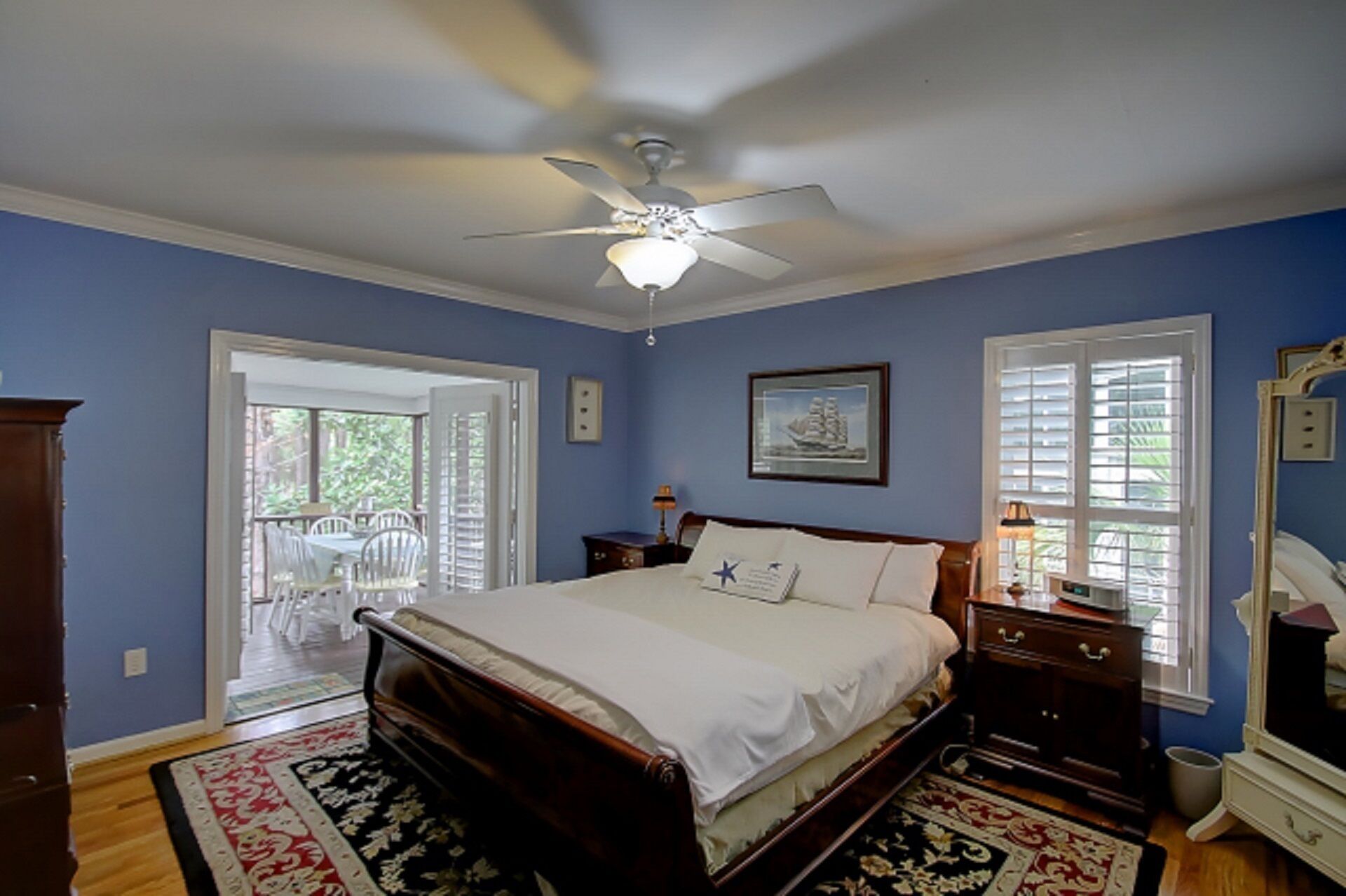 Une chambre à coucher comprenant un lit, un ventilateur de plafond, une fenêtre avec des stores, une commode et un miroir.