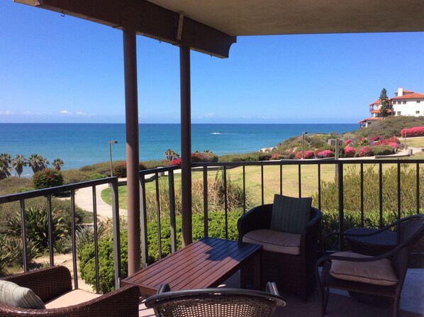 A balcony with wicker furniture, a wooden table, and a view of the ocean and a house.