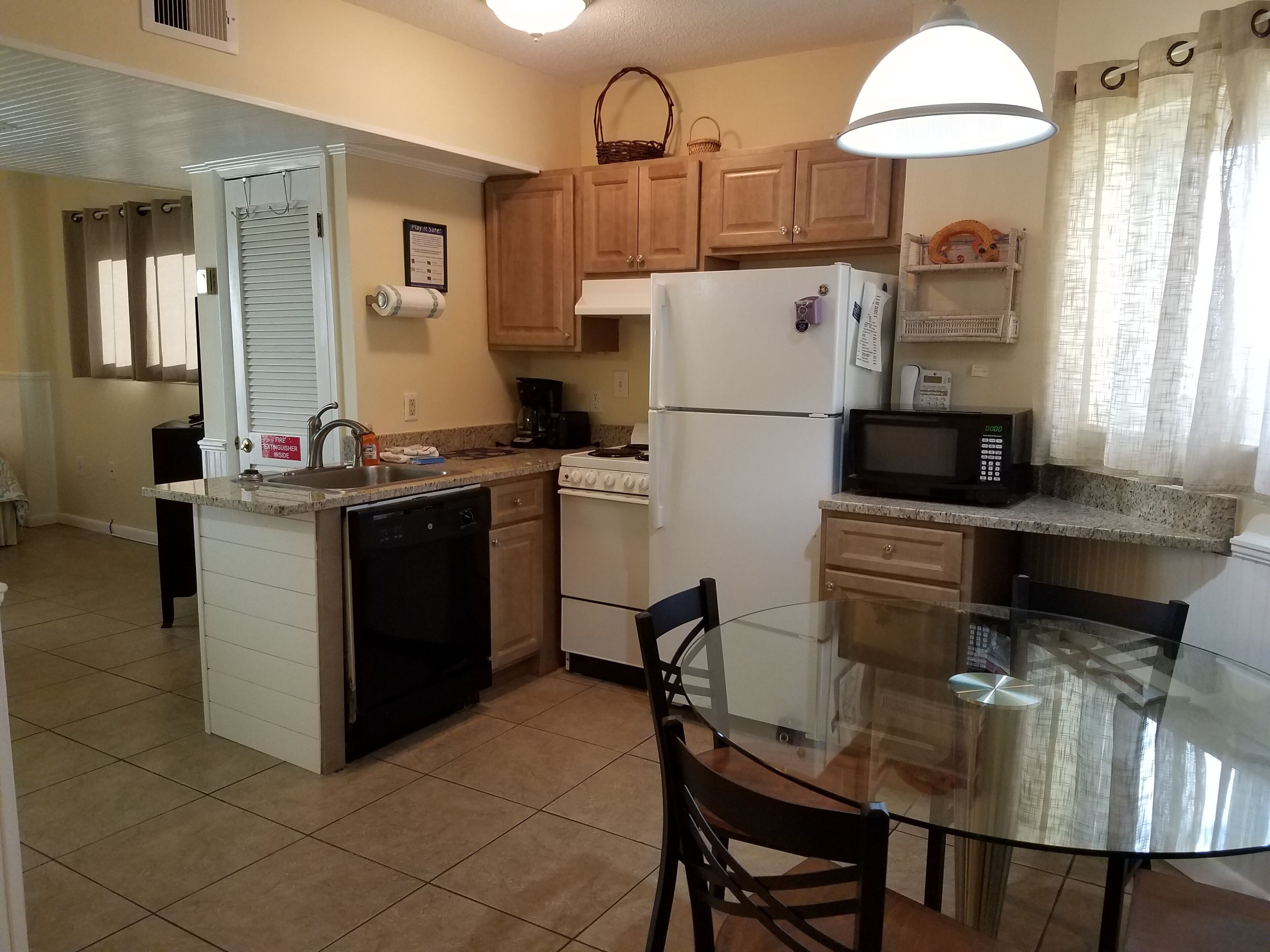Cocina compacta con lavavajillas, estufa, refrigerador y microondas. Hay una mesa de vidrio con sillas y una ventana con cortinas.