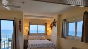 A bedroom with a bed, two windows, a ceiling fan, and a view of the sea.