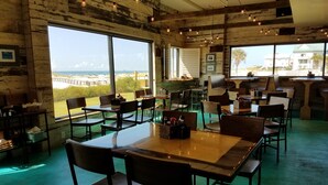 A restaurant with large windows offering a view of the beach and ocean. Tables and chairs are arranged indoors.