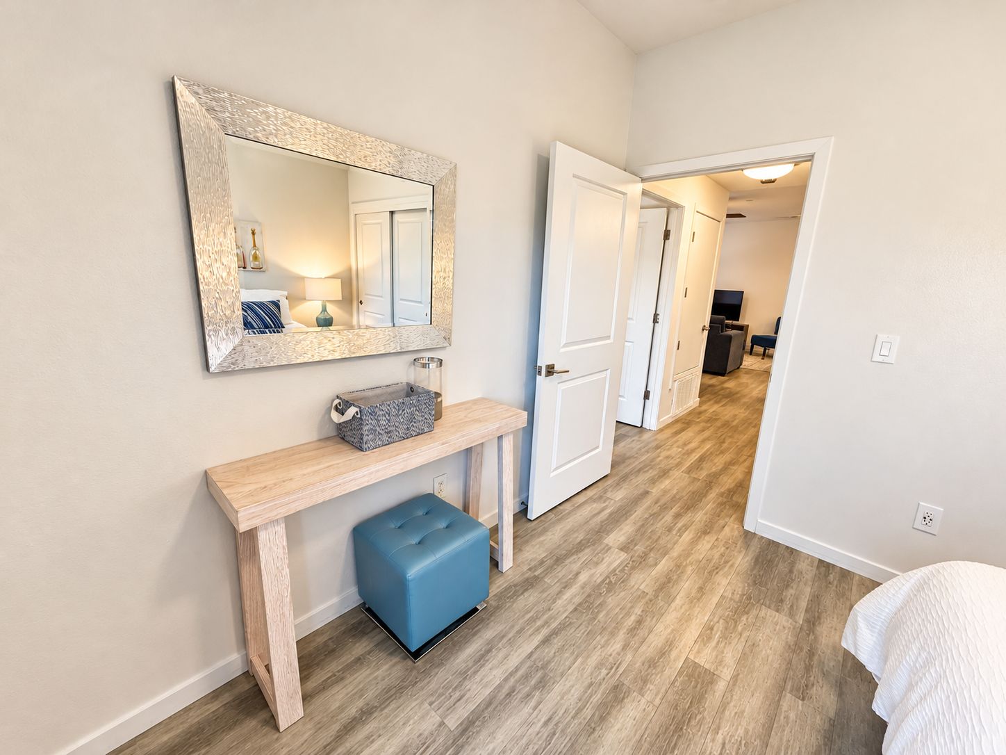 A hallway with a wooden bench, a blue ottoman, a mirror, and a door leading to another room.